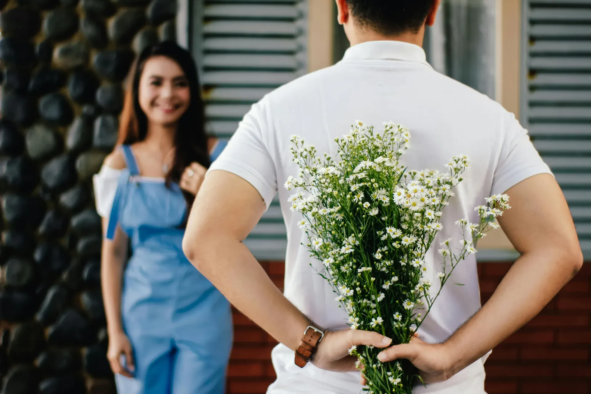 Person hiding a bouquet of white flowers behind their back while greeting a smiling partner at the door.