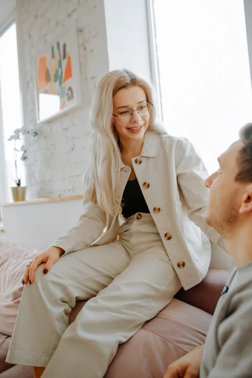 Woman in light clothing smiling gently while sitting and talking with a man indoors, representing reconnection and healthy relationship communication.