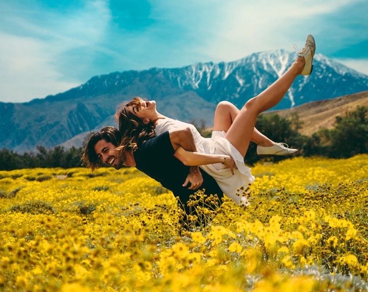 A laughing man gives a woman a piggyback ride in a vibrant field of yellow flowers.