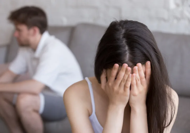 Woman covering her face after an argument while her partner sits in the background, representing insecurity and disconnection in a relationship.