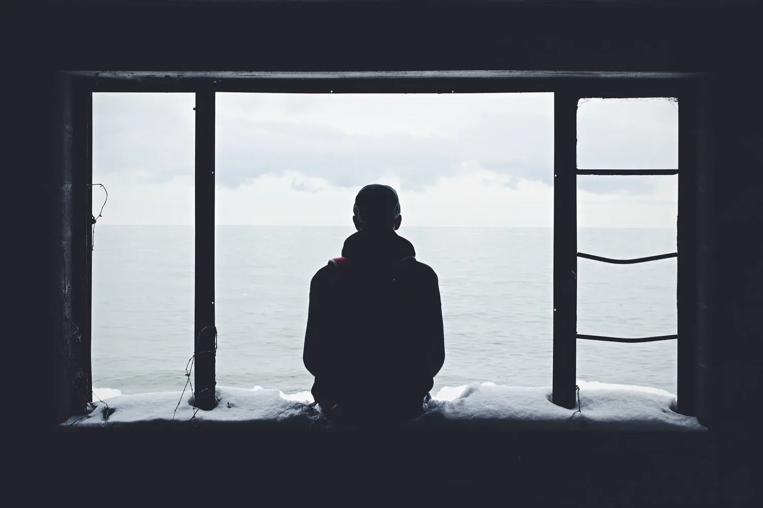 Person sitting alone by a window facing the sea, symbolizing solitude, reflection, and emotional resilience in love