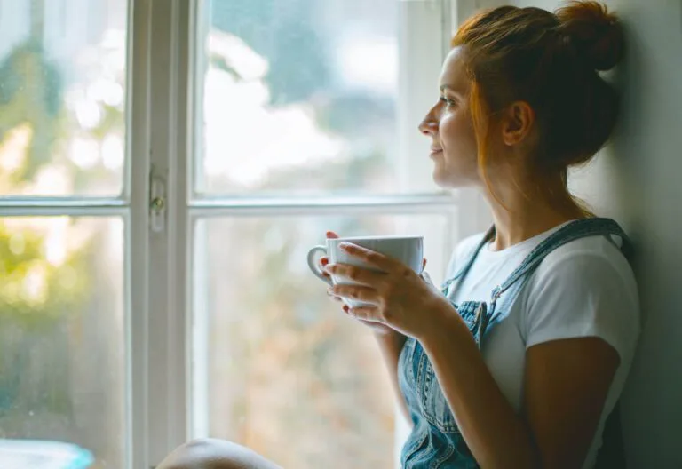 Woman holding a cup by a bright window, representing solitude, healing, self-care, and emotional peace after graduation.