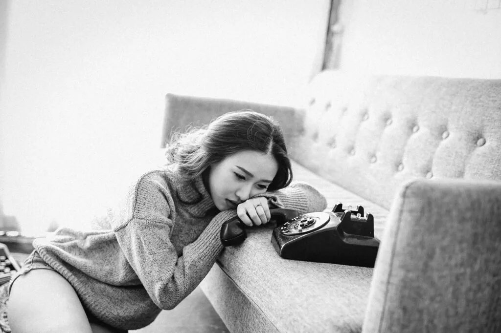 Black and white image of a woman leaning by a sofa beside a rotary phone, looking sad and emotionally drained.