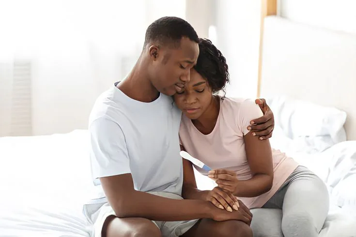 Couple sitting on a bed embracing while looking at a pregnancy test, symbolizing infertility, comfort, and relationship resilience.