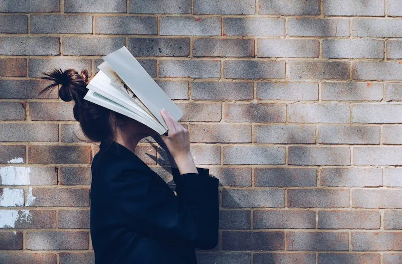 Person standing against a brick wall with an open book covering their face, representing discomfort, introspection, and personal growth.