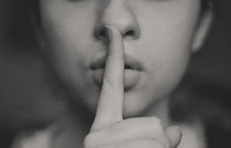 Close-up black-and-white image of a child making a shushing gesture with a finger to their lips.