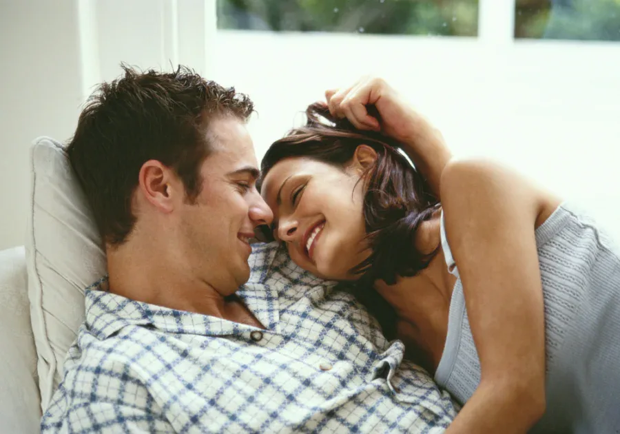 A smiling couple cuddling on a couch near a window, symbolizing tenderness, connection, and supportive relationships during major milestones.