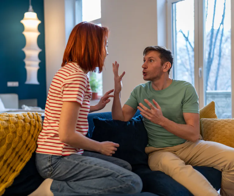 Casal tendo uma discussão intensa no sofá de casa, mostrando tensão no relacionamento, falhas de comunicação e conflito conjugal.