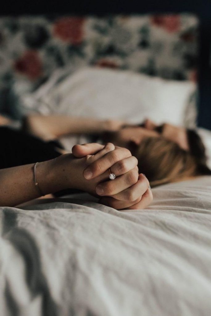 Couple holding hands in bed, one wearing a diamond ring.