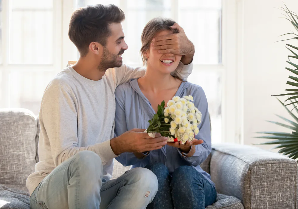 Man surprising a smiling woman with flowers on a sofa, illustrating affectionate gestures, thoughtful giving, and the emotional value of meaningful gifts.