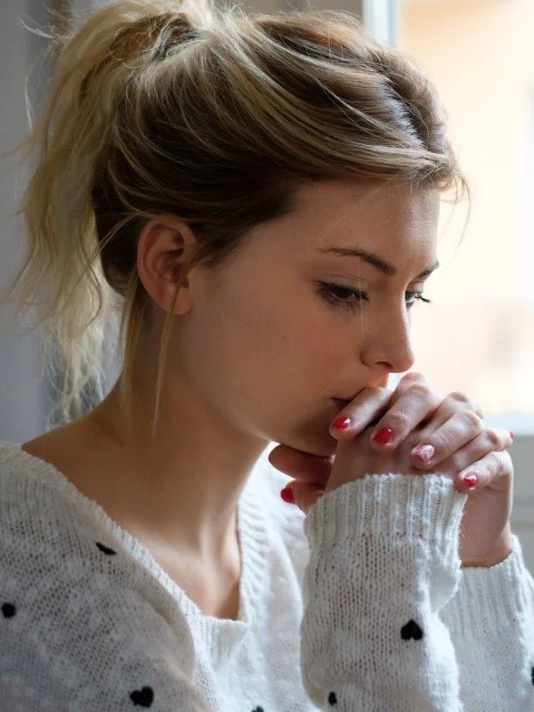A thoughtful young woman by a window with clasped hands, expressing reflection, worry, and emotional growth before a life milestone.