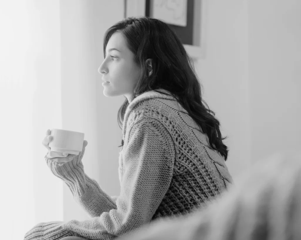 A woman in a sweater holding a cup and looking out a window, symbolizing healing after emotionally unavailable relationships.