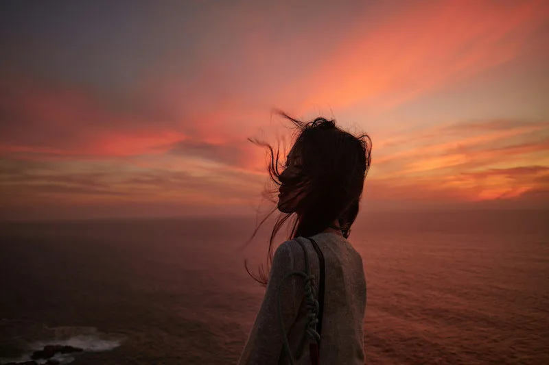 Woman silhouetted against a pink and orange sunset over the ocean, symbolizing hope, self-love, and a fresh New Year start.