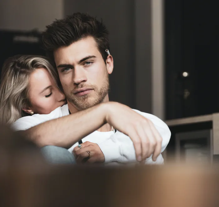 Woman hugging a serious man from behind indoors, expressing closeness mixed with emotional strain.