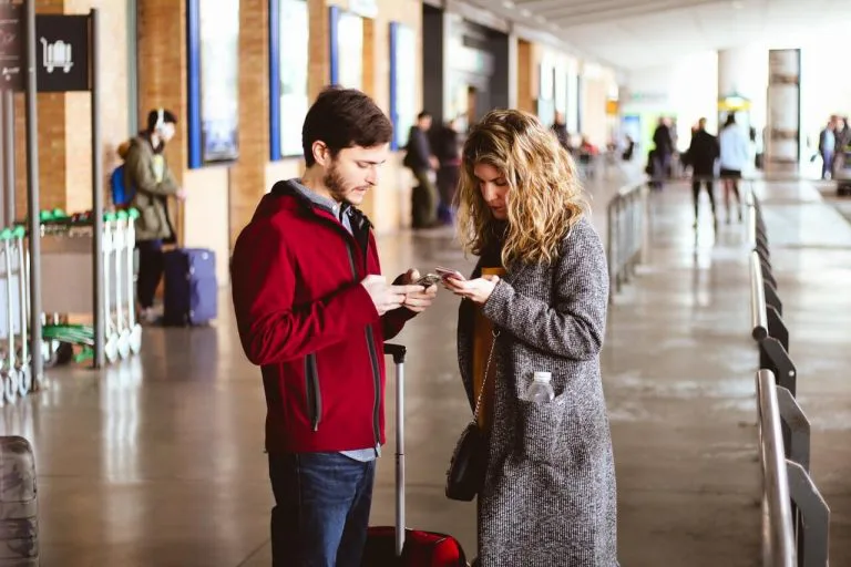 A man and woman standing in a transit terminal looking at their phones, symbolizing distraction, emotional distance, and modern relationship challenges.