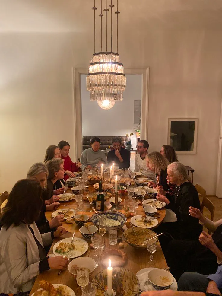 A group of family and friends gathered around a candlelit dinner table in a warm home setting.