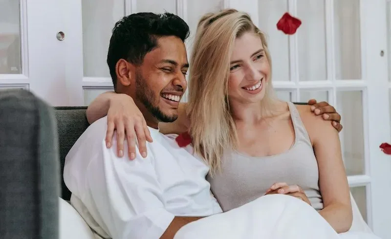 A happy couple sitting close together in bed, smiling and embracing in a bright intimate room.