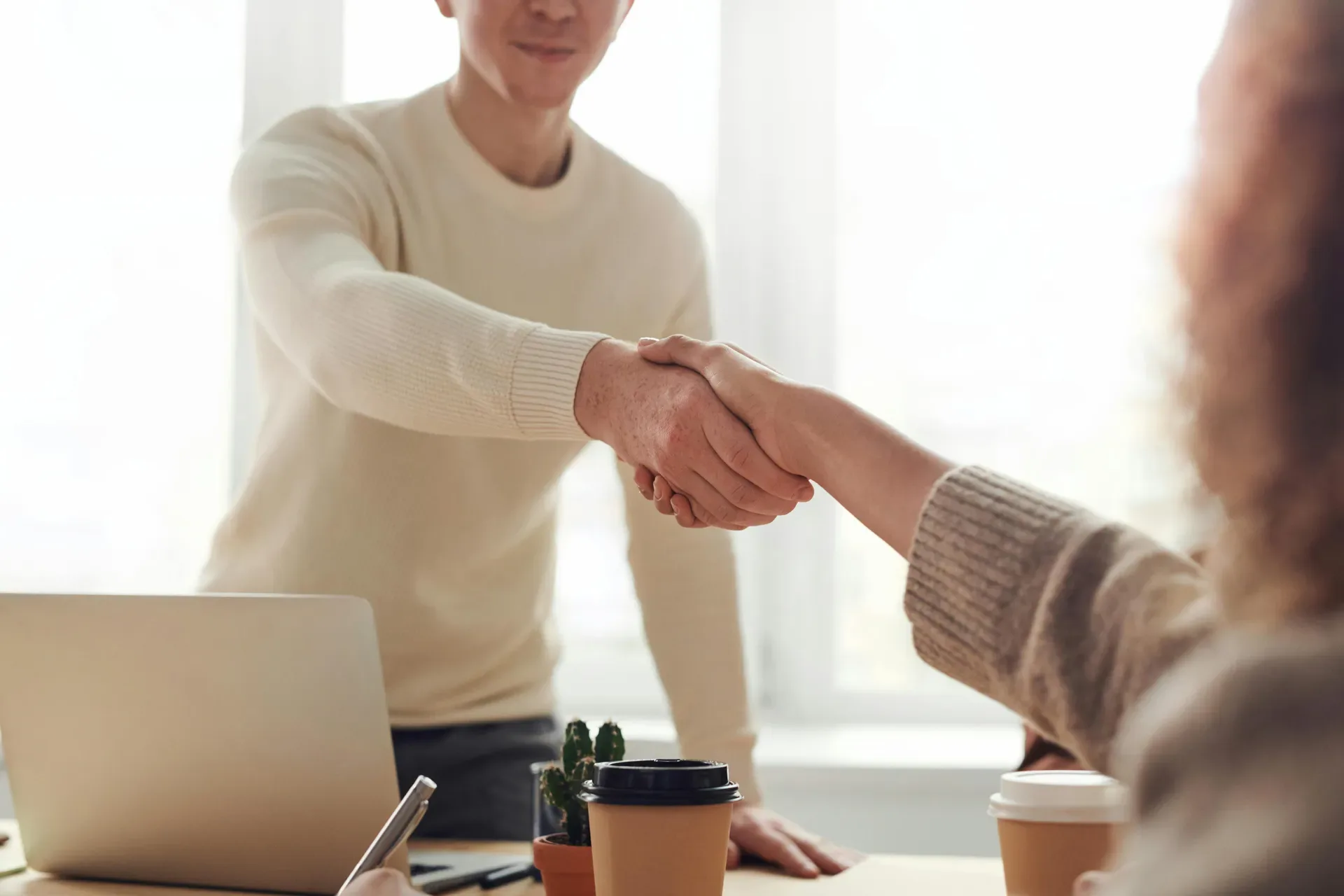 Two people shaking hands across a table near a laptop and coffee cups, representing reconciliation, commitment, and relationship reset