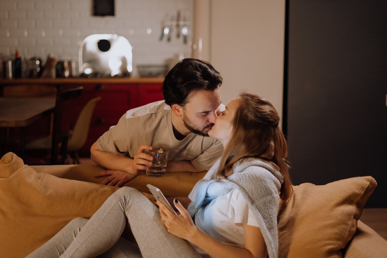 A man and woman sharing a tender kiss on a warm, dimly lit couch. The woman is holding a smartphone and the man holds a glass.
