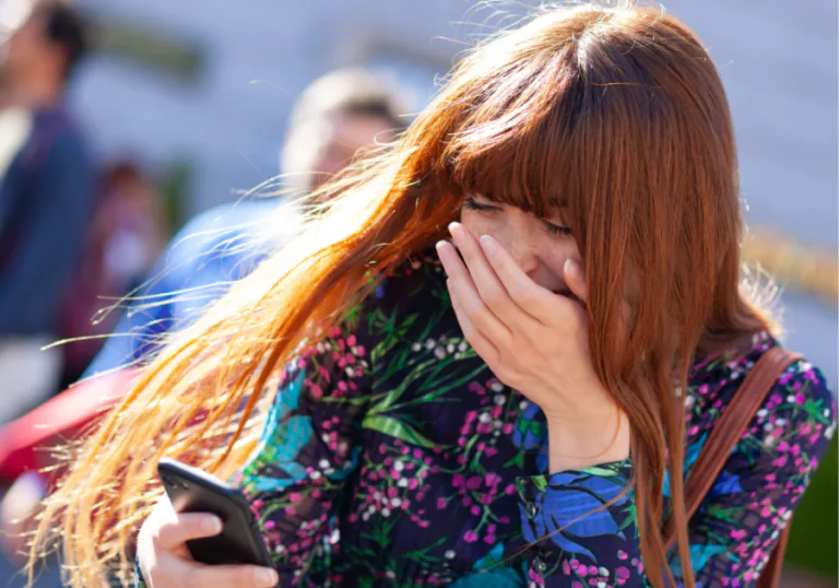 Mulher jovem ao ar livre, chocada e emocionada ao ler algo no celular.