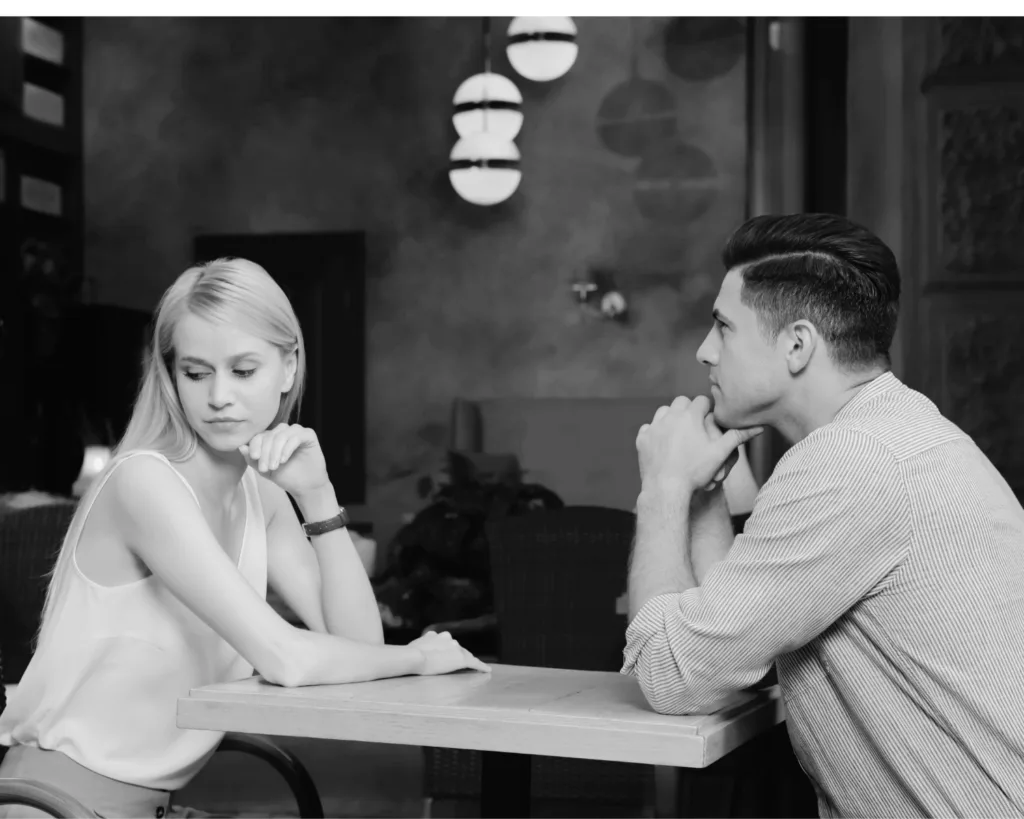 Black and white image of a couple sitting apart at a table, showing emotional distance and relationship tension.