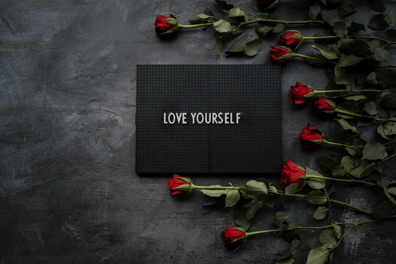 Black card with the words Love Yourself surrounded by red roses on a dark surface, symbolizing self-worth and romantic reflection.