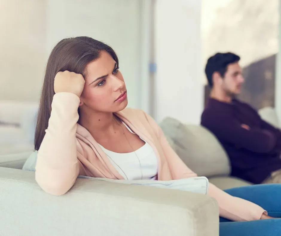 A thoughtful woman in the foreground and a distant man in the background on a couch, illustrating emotional disconnection in a couple.