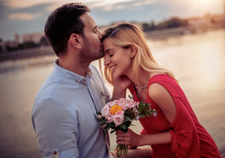 Couple embracing at sunset by the water, with the woman holding a small bouquet of flowers.