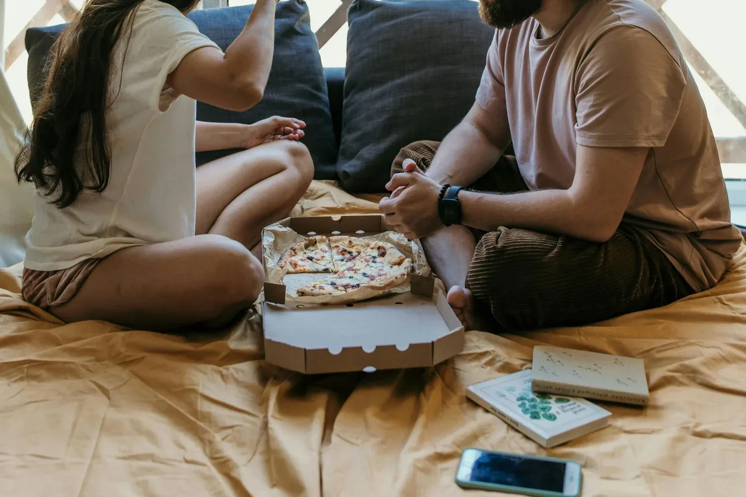 Couple sitting on a bed sharing pizza and conversation in a relaxed, intimate home setting.