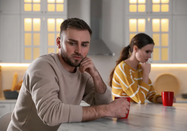 A sad couple sitting apart in a kitchen, each holding a mug and looking troubled after a conflict.