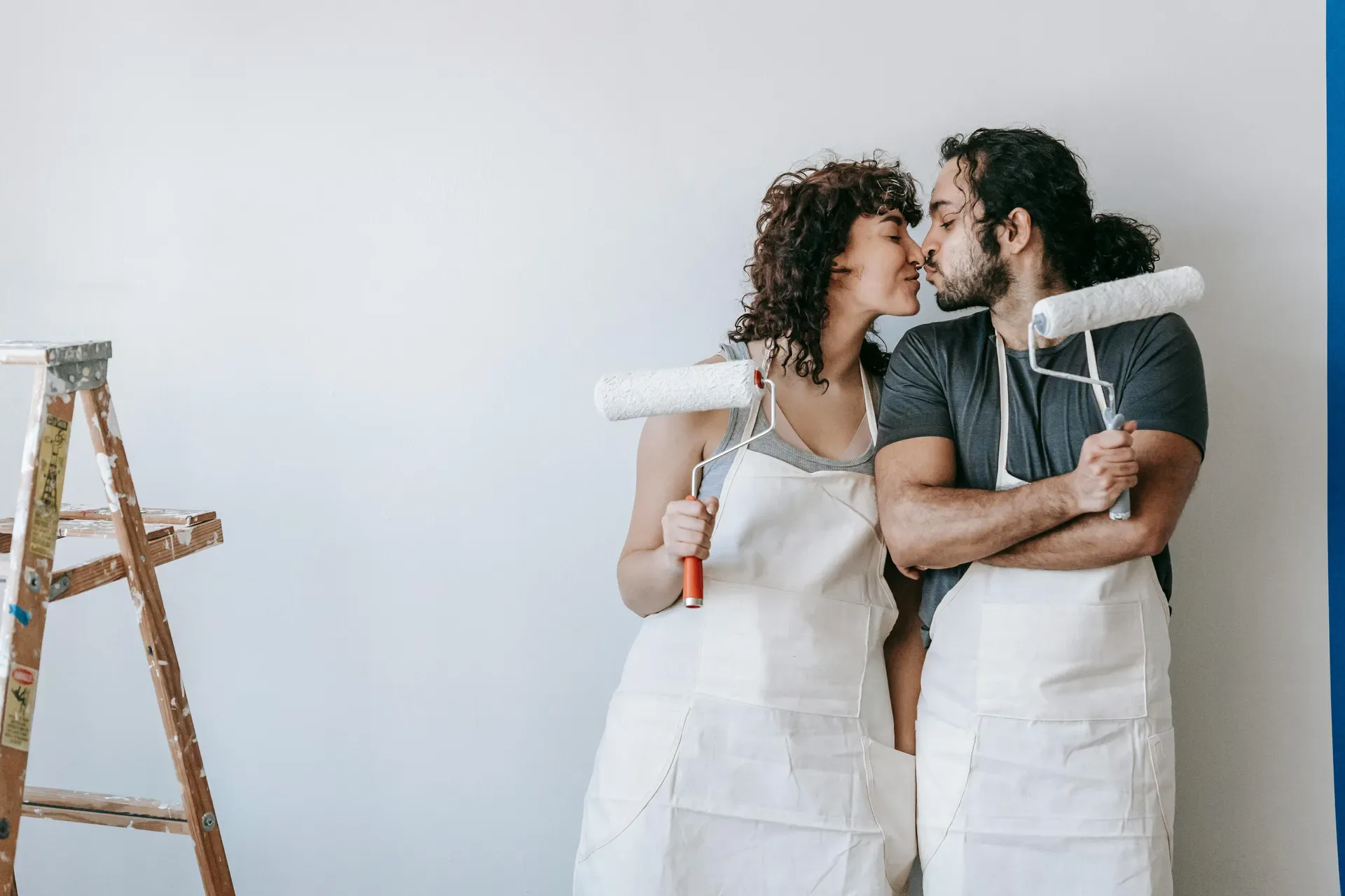 Couple in white aprons holding paint rollers and kissing while renovating a room together.