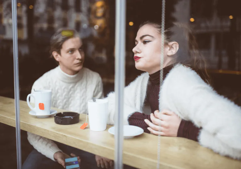 A tense café scene with a woman looking away and a man watching her, suggesting emotional distance and unresolved feelings.