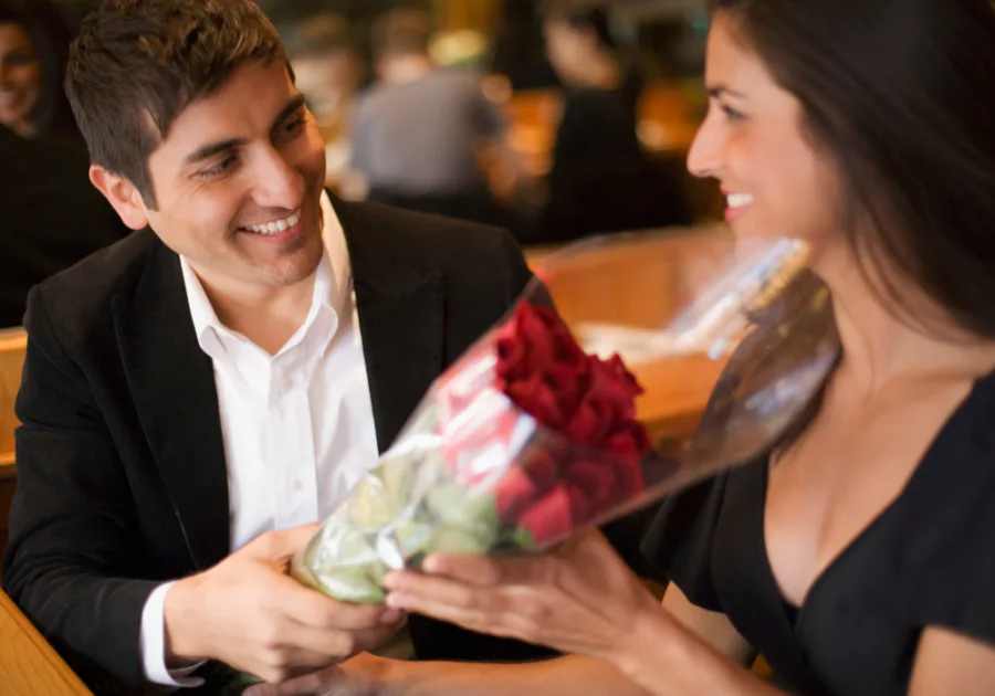A man gifting red roses to a smiling woman across a restaurant table in a romantic setting.