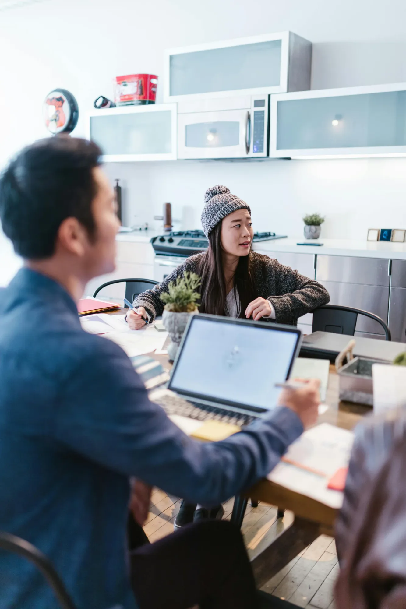 Two people talking at a table with laptops and papers in a bright kitchen, representing busy schedules, communication, and relationship stress.