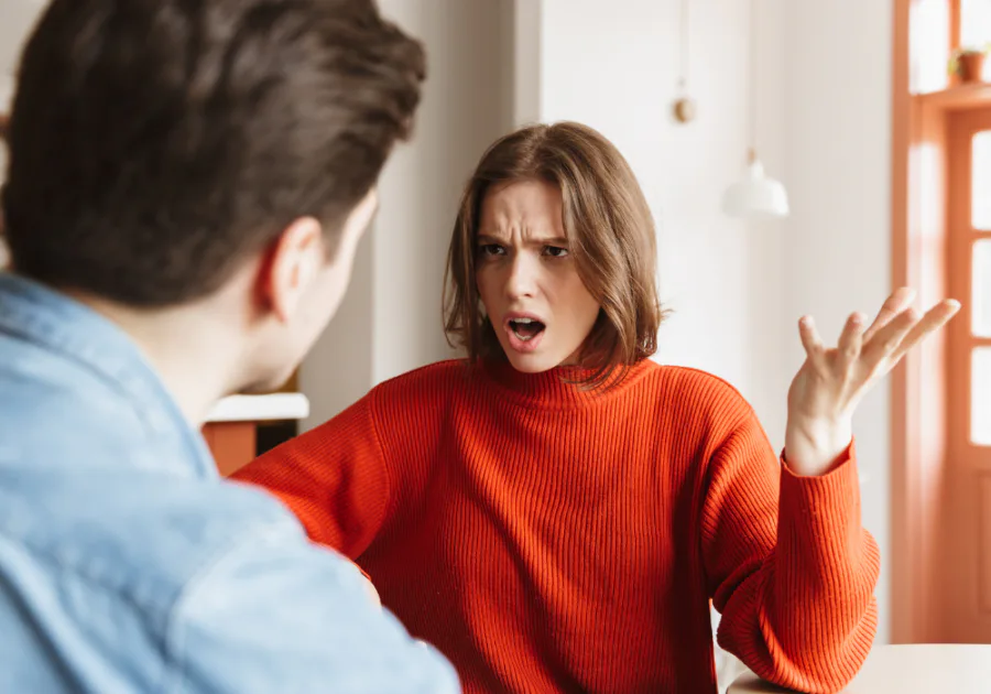 Woman in a red sweater arguing indoors with a partner, expressing frustration and emotional overwhelm in a strained relationship.