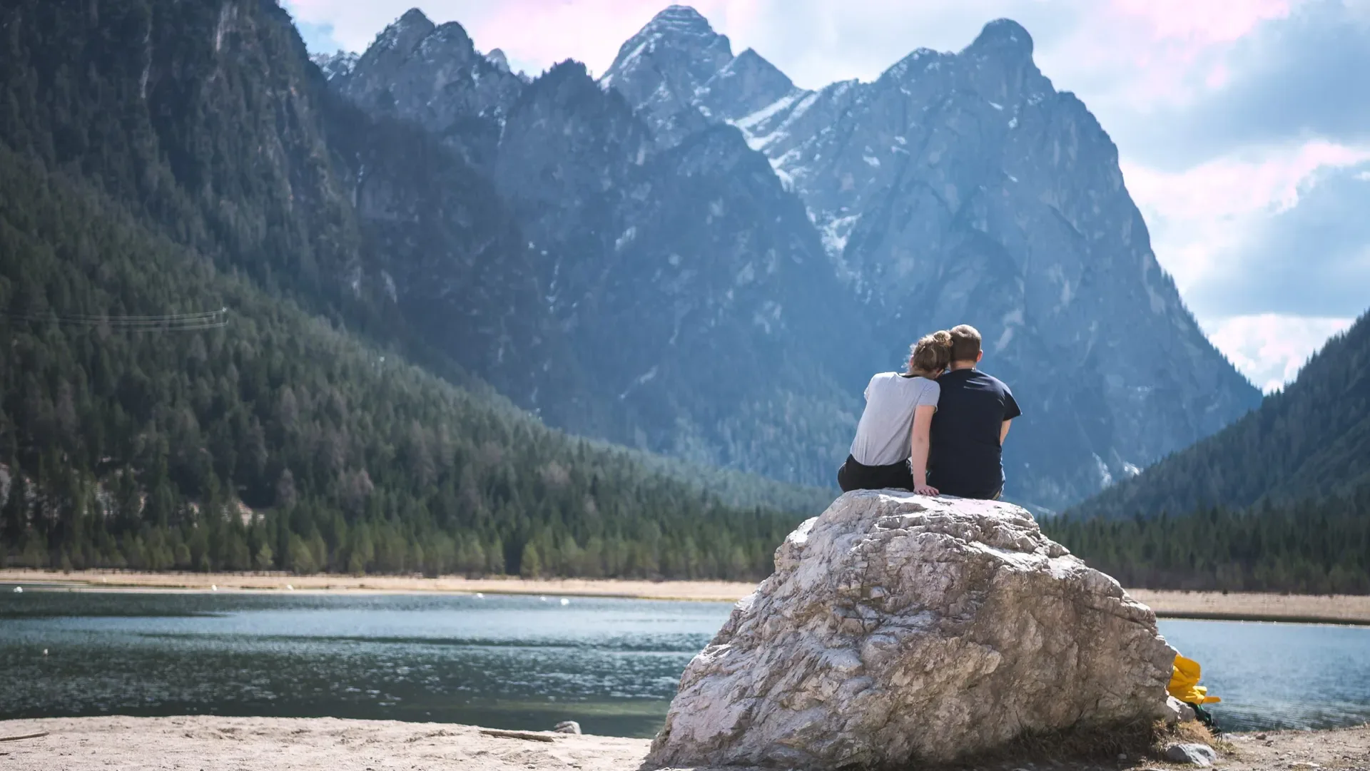 Couple sitting on a rock by a lake with mountains in the background, symbolizing reflection, achievement, and the journey ahead.