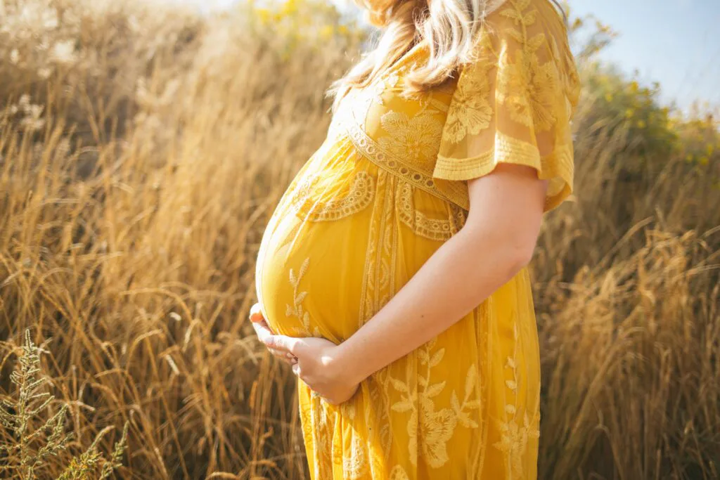 Pregnant woman in a yellow dress holding her baby bump in a sunny field, symbolizing hope, vulnerability, and a new chapter ahead.