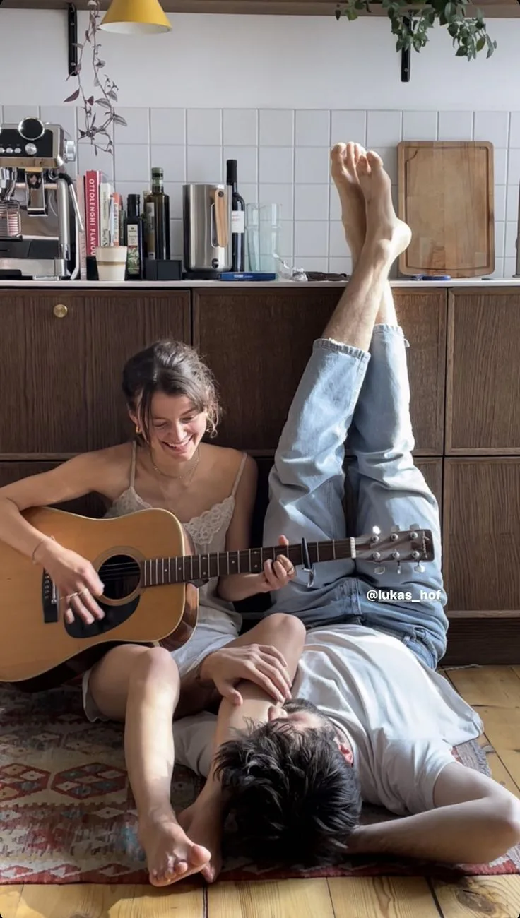 A woman playing guitar while sitting on the floor beside a relaxed partner in a cozy kitchen, showing playful romantic intimacy.