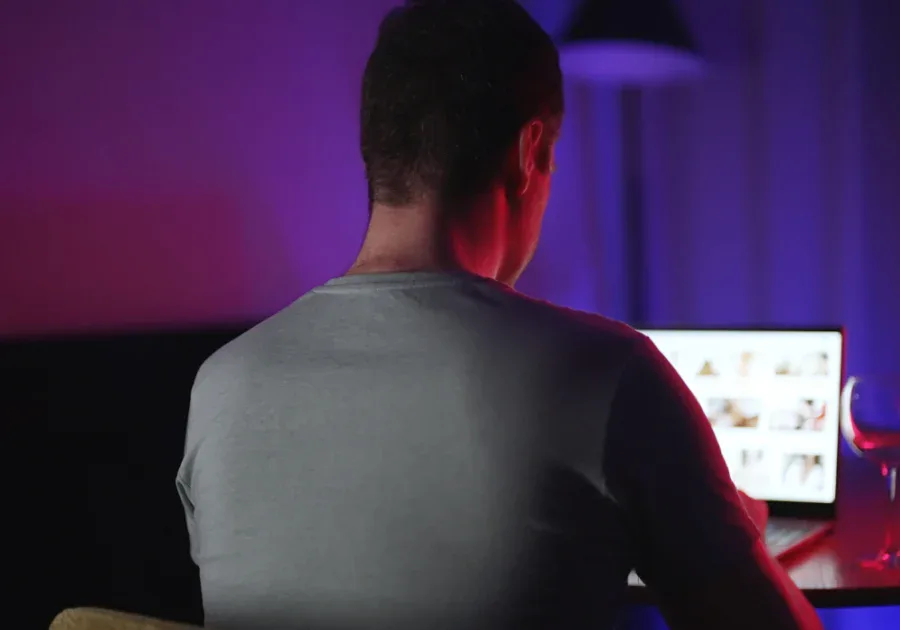 Homem sentado sozinho em frente a um computador em um quarto escuro iluminado por luz roxa e azul, representando isolamento digital nos relacionamentos