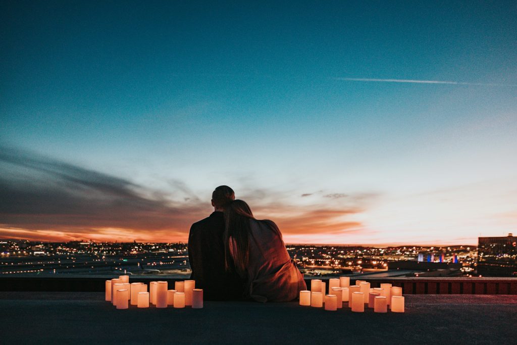 Sepasang pasangan duduk bersama saat matahari terbenam dikelilingi lilin dengan pemandangan bandar, melambangkan romantik dan keintiman ulang tahun.