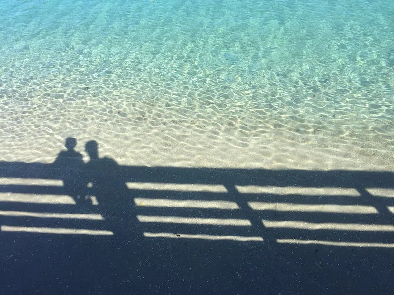 Shadows of two people on a bridge cast over bright, clear blue water below.
